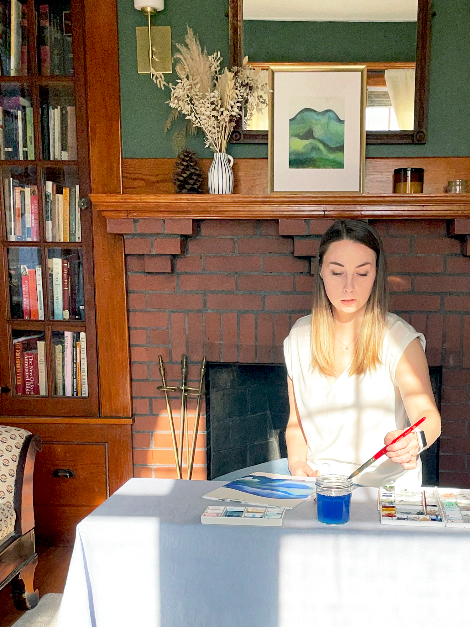 Amity Buchanan is sitting at a desk painting with watercolors. There is a fireplace, bookcase, and green walls behind here. The painting is not visible but a jar of bright blue water sits on the table. Afternoon shadows fall across the image. 